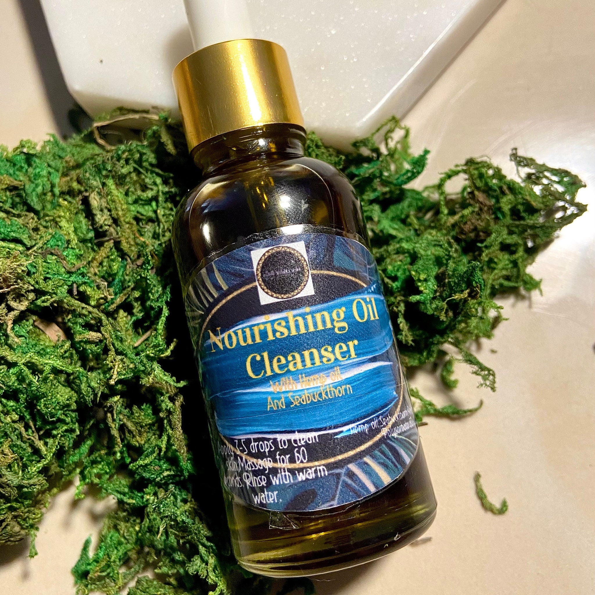 Facial Oil Cleanser bottle with gold cap, blue label, and green moss background