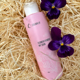 Face powder product in pink bottle with floral design, purple flowers, and straw background