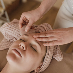 Gua sha tool being used on face, light pink stone, light skin, towel wrap