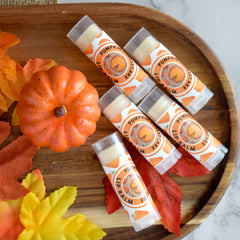 Lip balm tubes with orange and white labels, arranged on a wooden tray with fall leaves and pumpkin decoration