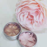 Eyeshadow makeup, two metal tins with pink and brown powder, next to pink flower petals