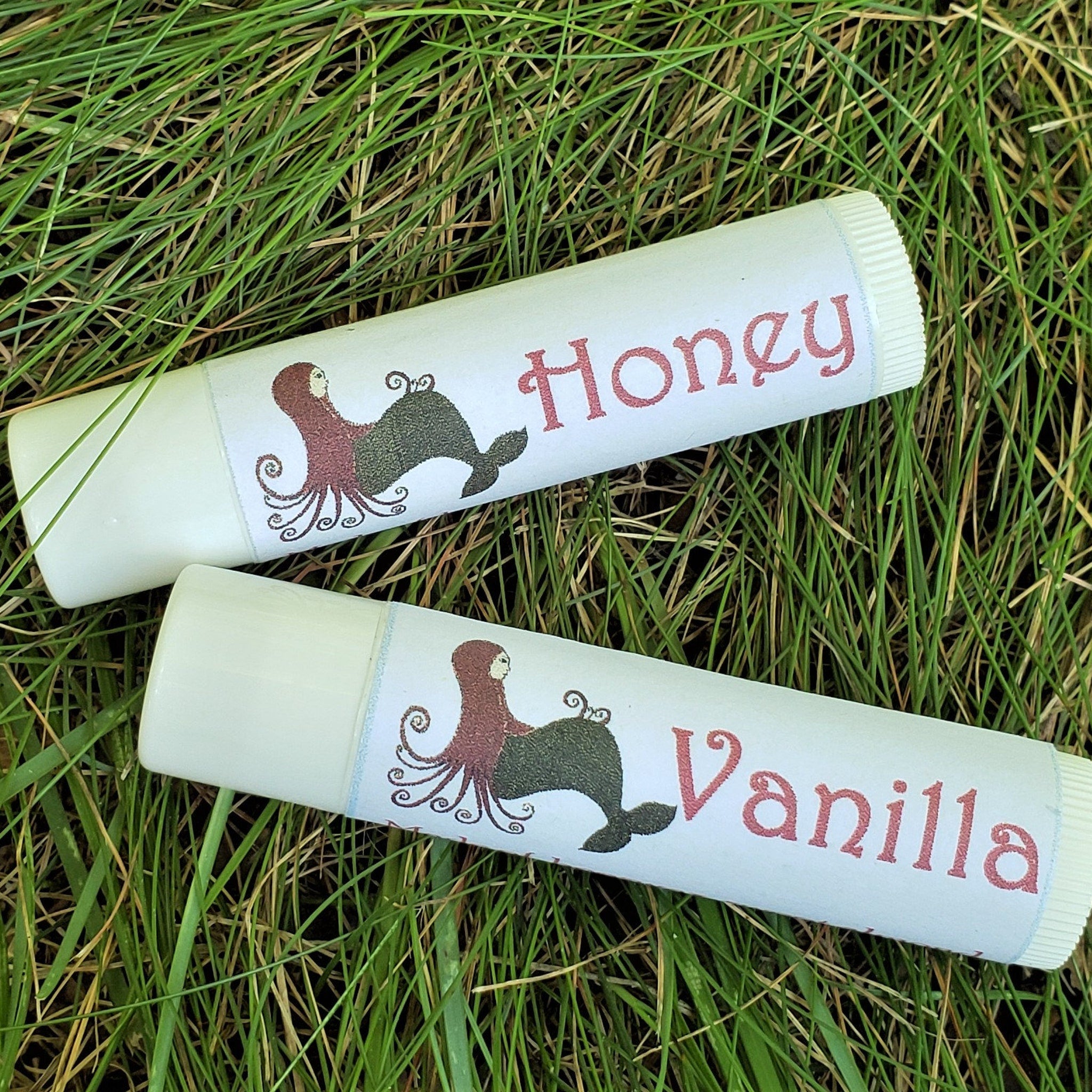 Lip balms, white tubes with red "Honey" and "Vanilla" text, Meadow Mermaids brand, on grass