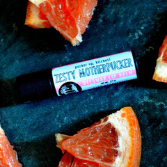 Lip balm tube with black, pink, and white label, and grapefruit slices on dark blue surface