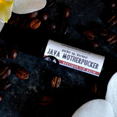 Lip balm tube with white and red label, surrounded by brown coffee beans and white flower petals, Java Motherpucker text
