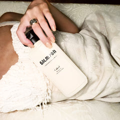 Facial cleanser bottle, cream-colored, black cap, held by hand with ring, Caked brand visible