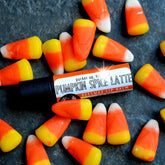 Lip balm tube with black base, orange and white label, surrounded by candy corn on textured surface