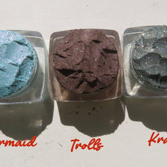 Side view of six vibrant loose powder eyeshadows in monster-themed shades with shimmer finish, displayed in round jars