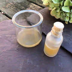 Glass jar with yellow liquid, small glass bottle with cork, on dark stone surface.