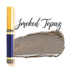 Eyeshadow swatch in grey-brown color with gold and navy tube, "Smoked Topaz" text visible