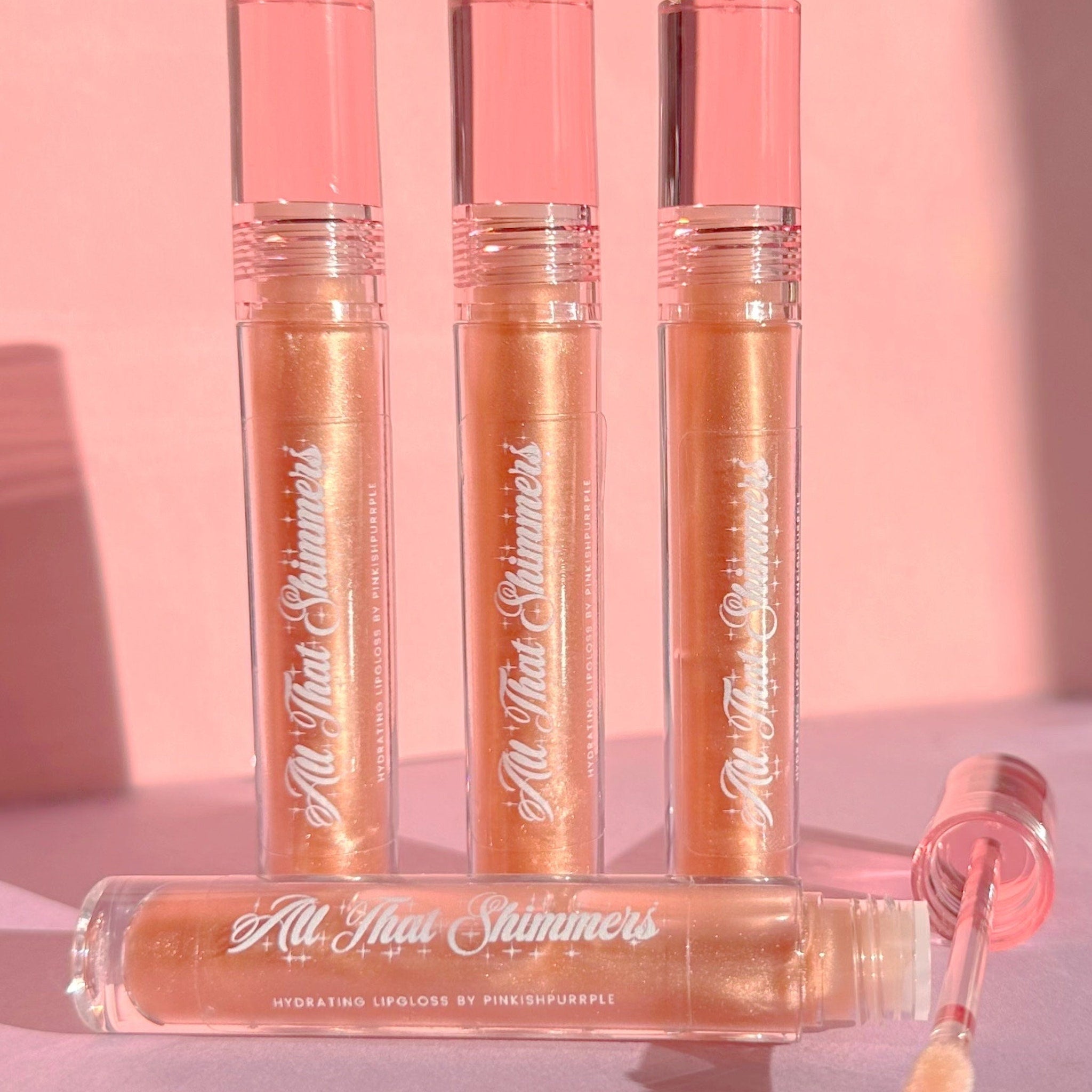 Lip gloss tubes in peach and pink with "All That Shimmers" text, clear applicator, PinkishPurrple brand