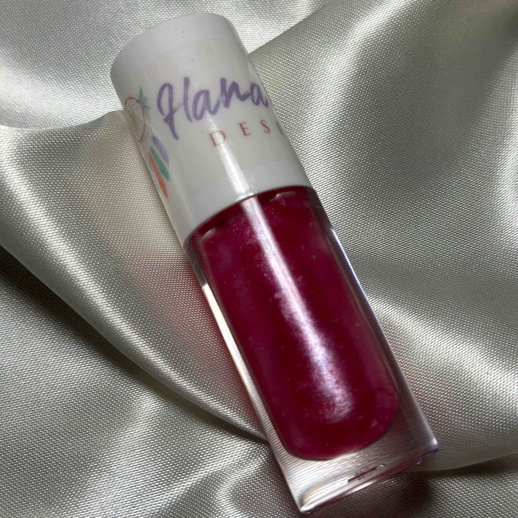 Lip gloss tube with red gloss, white cap, and "Hana Design" text on white satin fabric background