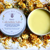 Calendula Salve, yellow salve in metal tin, surrounded by dried orange flower petals, Zuzu's brand visible