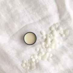 Lip balm in black container with light yellow balm on white linen background and white wax beads