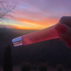Lip gloss tube in pink with silver cap, held by hand, with "Vainqueur by Victoria" text
