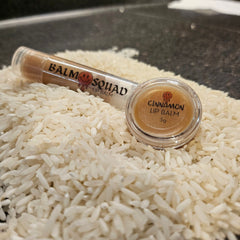 Lip balm with brown tint, clear tube, and "BALM SQUAD" text, on white rice grains.