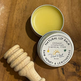 Lip balm in silver tin with yellow balm, wooden honey dipper, The Based Kitchen label on wood surface