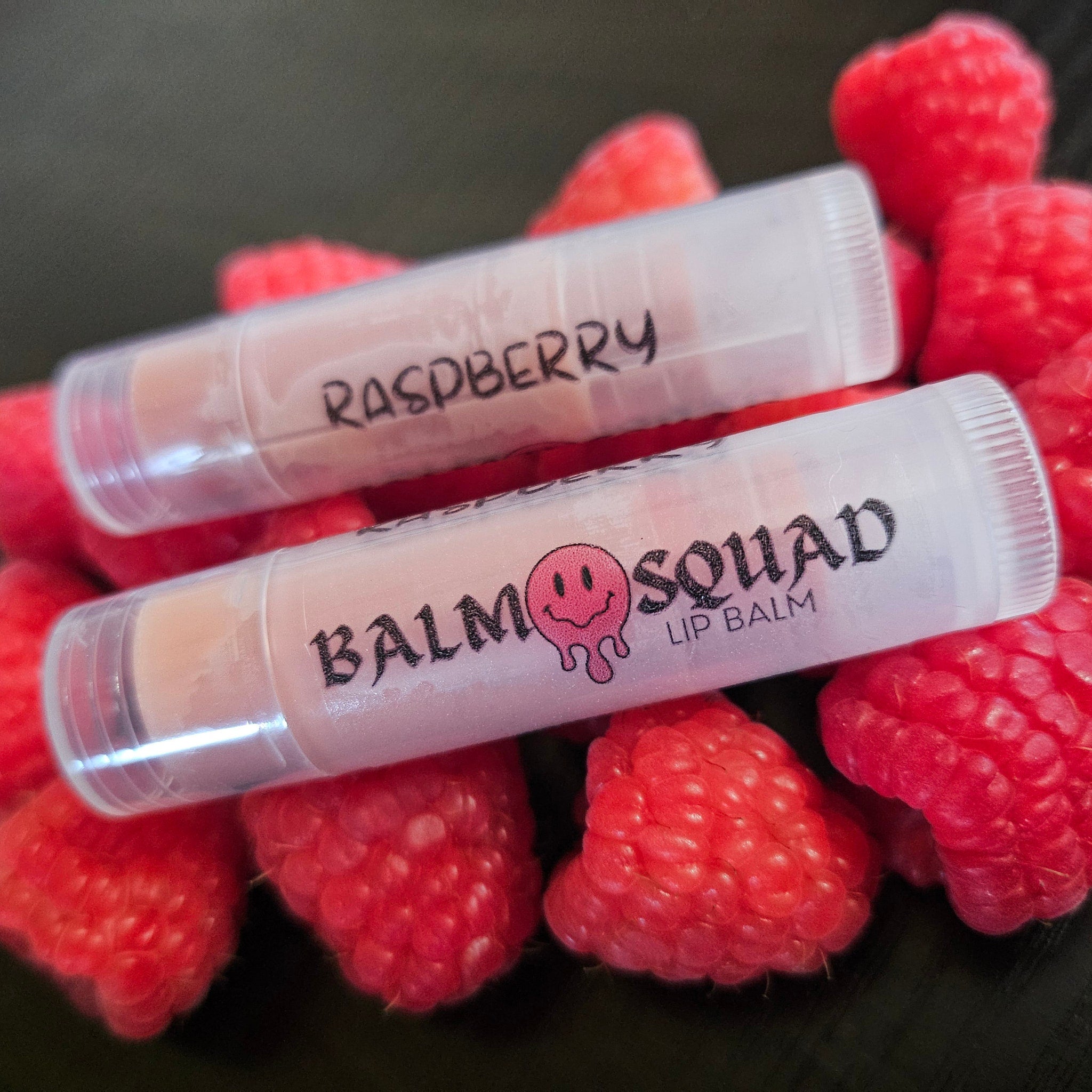 Lip balms, clear tubes with pink balm, "Raspberry" and "Balm Squad" text, surrounded by red raspberries