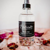 Toner spray bottle, clear glass, black label with "Potions" text, on pink cloth with rose petals