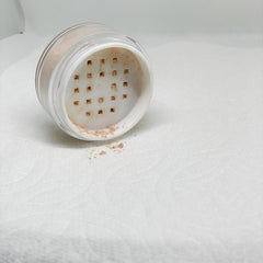 Loose powder makeup in clear plastic container with beige powder, spilled on white textured surface
