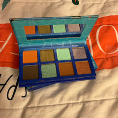 Eyeshadow palette with blue, teal, and orange packaging, featuring various colored eyeshadows, and brand name "Little Moonchild Goods" visible.