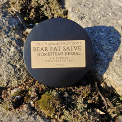 Bear fat salve, black metal tin, Homestead Herbal label, on mossy rock surface