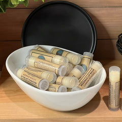 Lip balm tubes in clear plastic with tan labels, in white bowl, "Beeswax Lip Balm" text visible