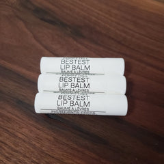 Lip balm tubes, white with black text, stacked on wood surface, Poppy Hill Bestest brand visible