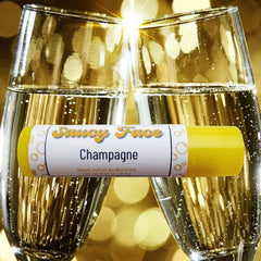 Lip balm with yellow tube, "Saucy Face Champagne" label, in front of champagne glasses
