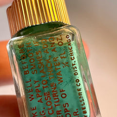 Vintage green eyeliner in glass bottle with golden spire lid displayed beside retro makeup accessories on velvet tray