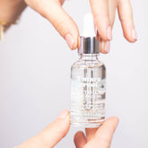 Face oil bottle held by hands, clear glass, silver cap, L'Etoile text visible