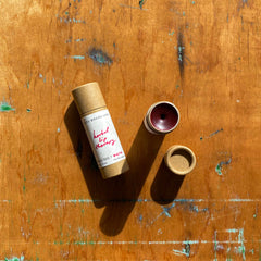 Lip balm tube with brown paper packaging, deep red tint, and open lid on wooden surface