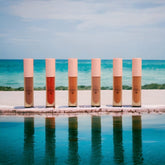Lip gloss tubes with pink caps, various shades, and VKC logo on a white surface by water