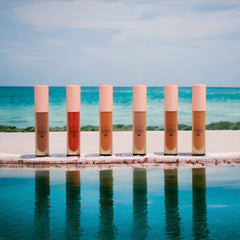 Lip gloss tubes with pink caps, various shades, and VKC logo on a white surface by water