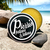 Facial Cleanser balm with yellow color, silver tin, and Pickle's logo on black lid on sand