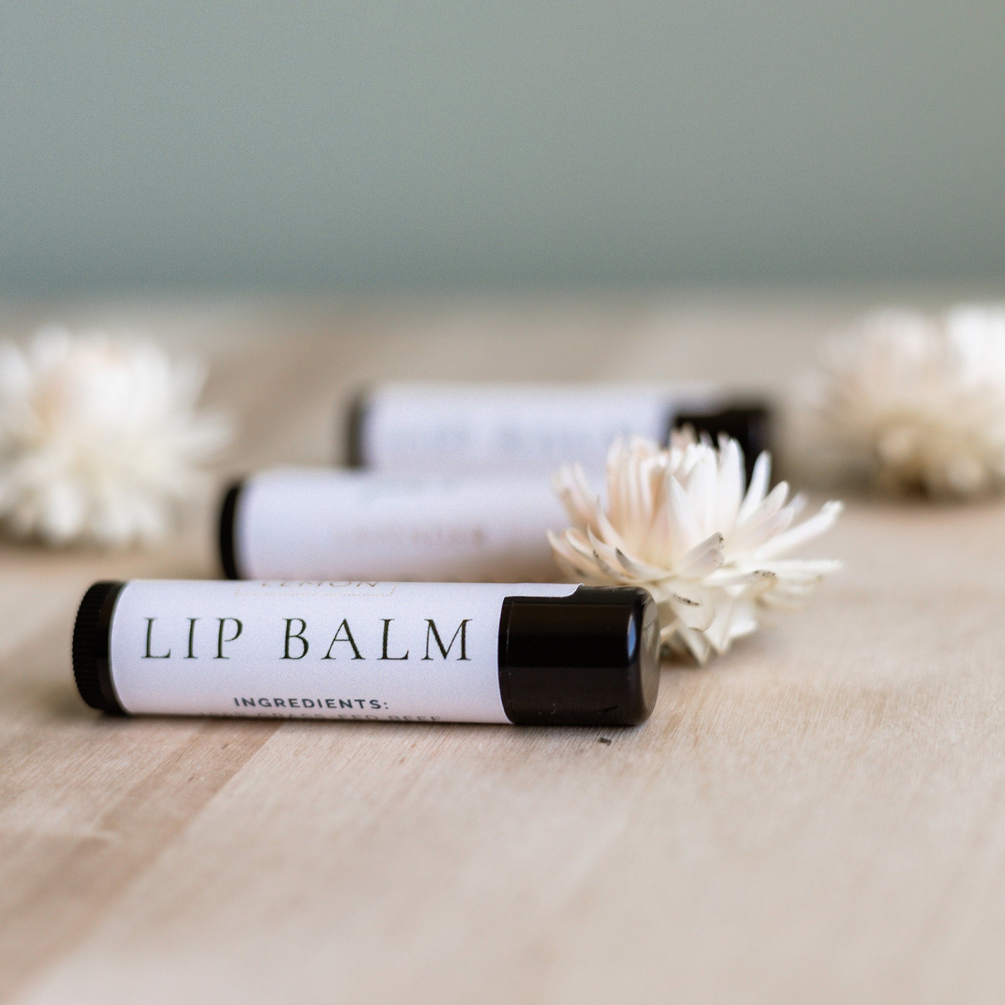 Lip balm tubes with white labels and black caps on wooden surface with flowers