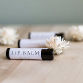 Lip balm tubes with white labels and black caps on wooden surface with flowers