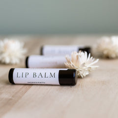Lip balm tubes with white labels and black caps on wooden surface with flowers