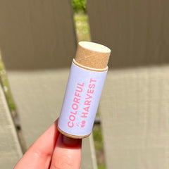 Lip balm tube with light pink and brown colors, and "Colorful Harvest" text label, held by hand