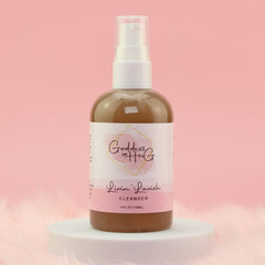 Cleanser bottle with white pump, brown liquid, and pink label with "Goddess in Her G" text on a white pedestal