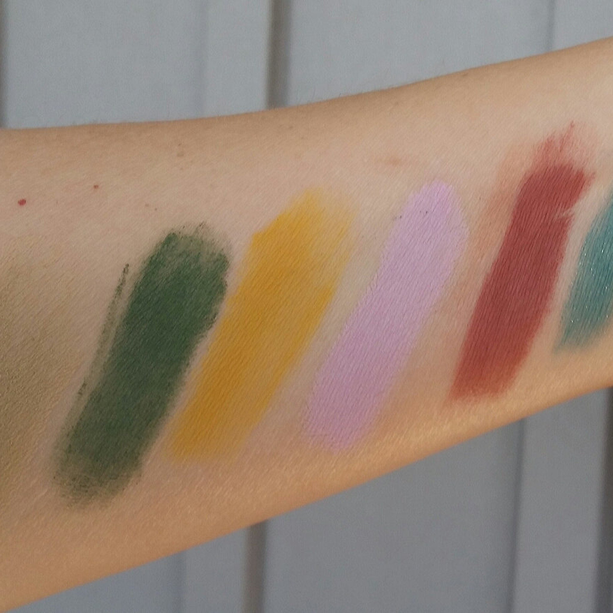 Eyeshadow swatches, various colors including green, yellow, pink, and red, applied on arm for color comparison