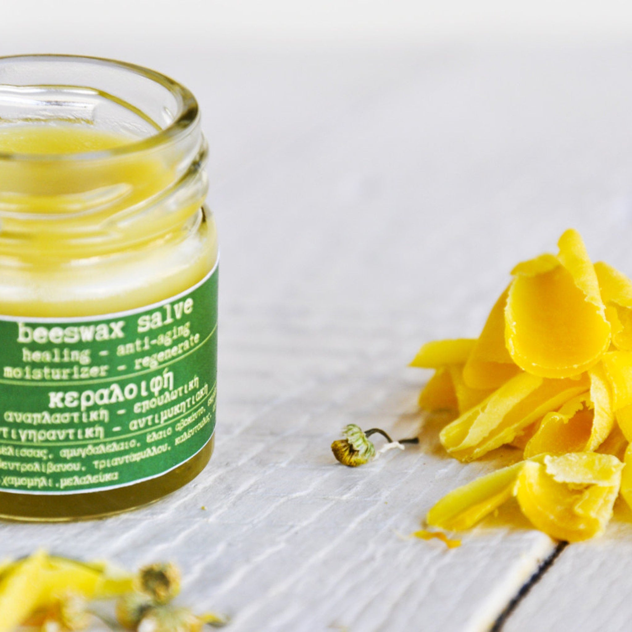 Beeswax salve in a clear glass jar with green label, yellow salve, and yellow wax flakes on white wood