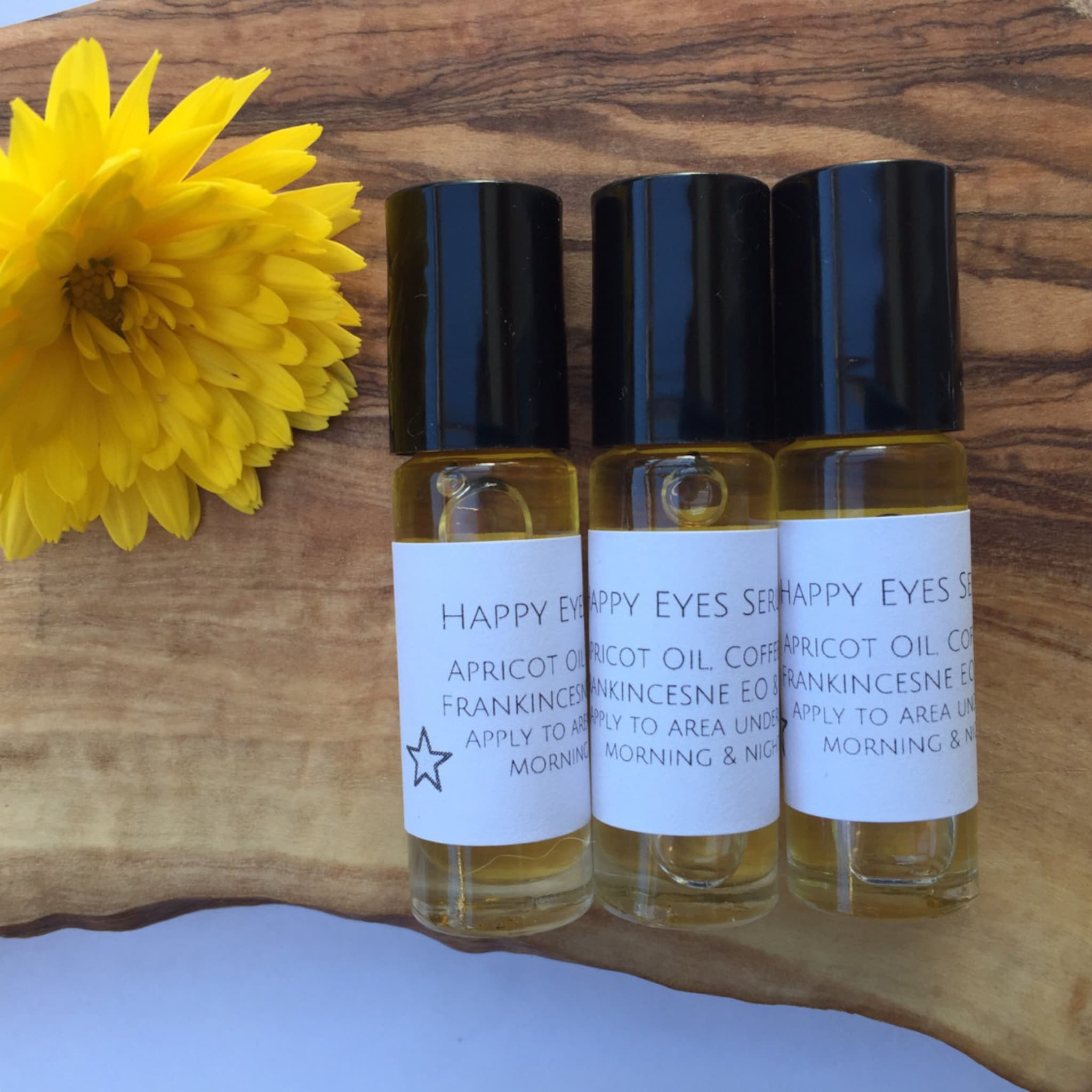 Eye serum bottles with black caps, clear liquid, and white labels on a wood surface with a yellow flower