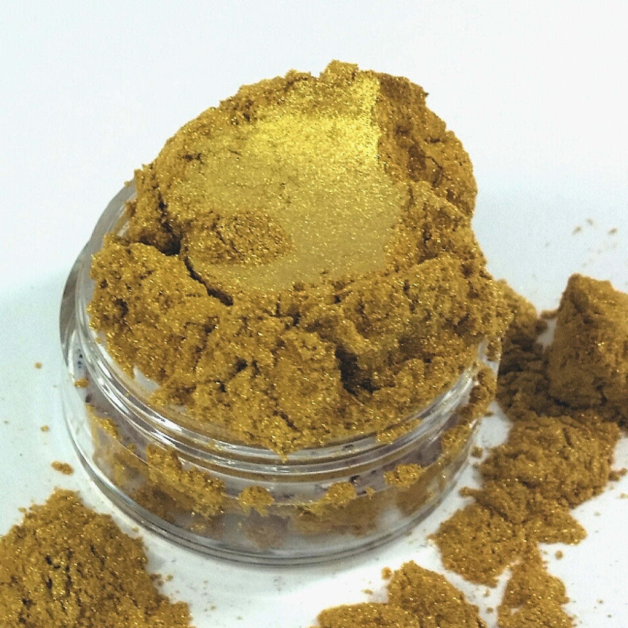 Gold pigment powder in a clear jar, with loose powder on a white surface, fine texture