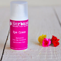 Eye cream bottle with pink label, white cap, and Herban brand text, on white wood surface with flowers