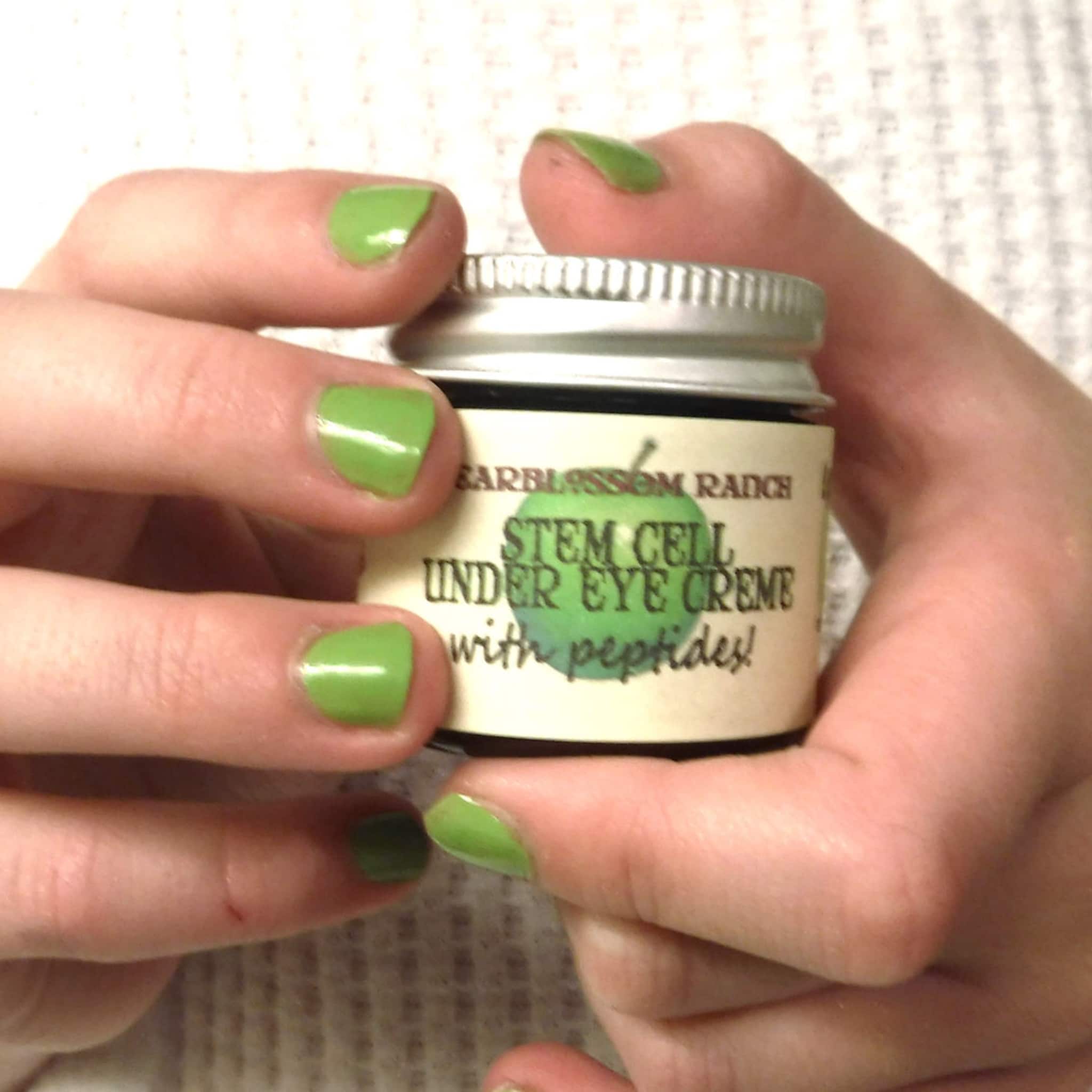 Under eye creme jar held in hands with green painted nails, metallic lid, and product label visible