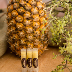 Lip balm tubes with clear plastic, yellow balm, and Pina Colada label, near pineapple