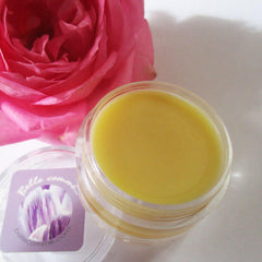 Yellow balm in clear plastic jar with Bellecomme label and pink flower in background
