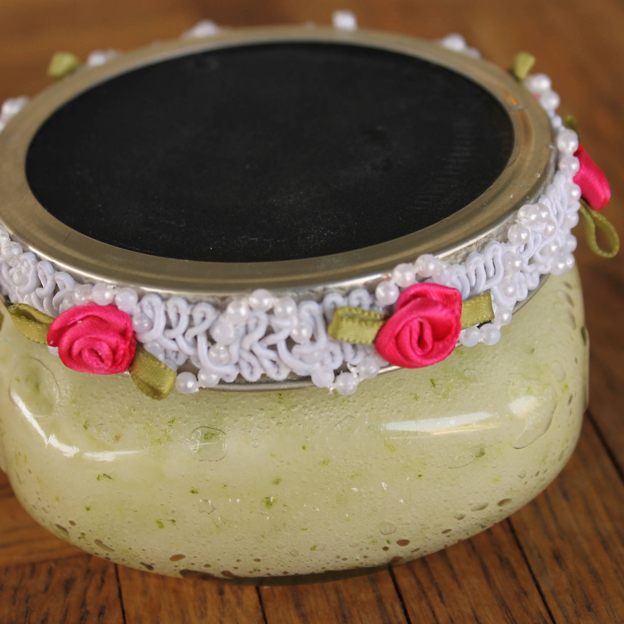 Glass jar with black lid, white lace trim, pink roses and pearls, on wooden surface
