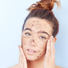 Facial scrub application, brown speckled paste on face, woman with hands touching face, light background
