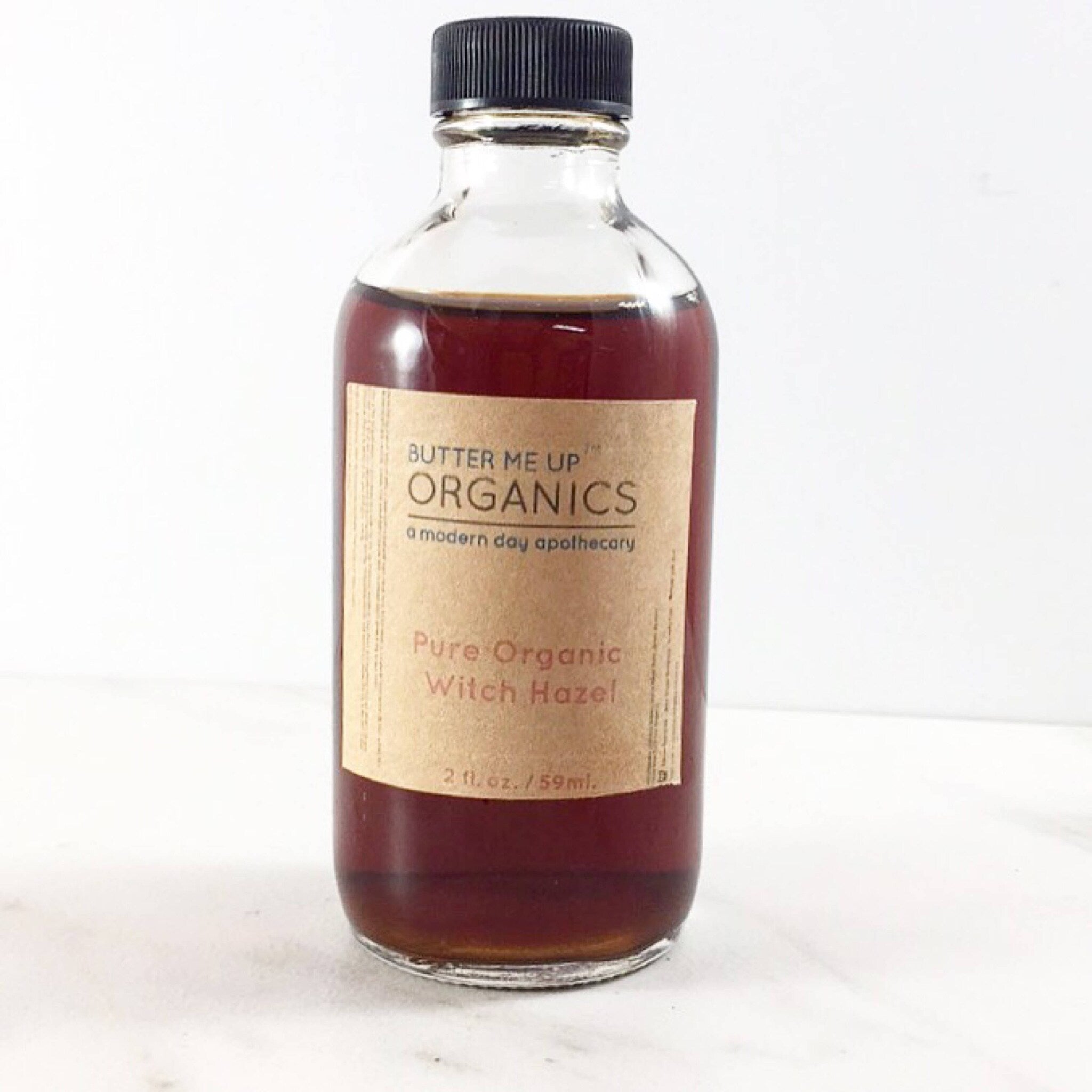 Witch hazel bottle with brown liquid, black cap, and paper label from Butter Me Up Organics